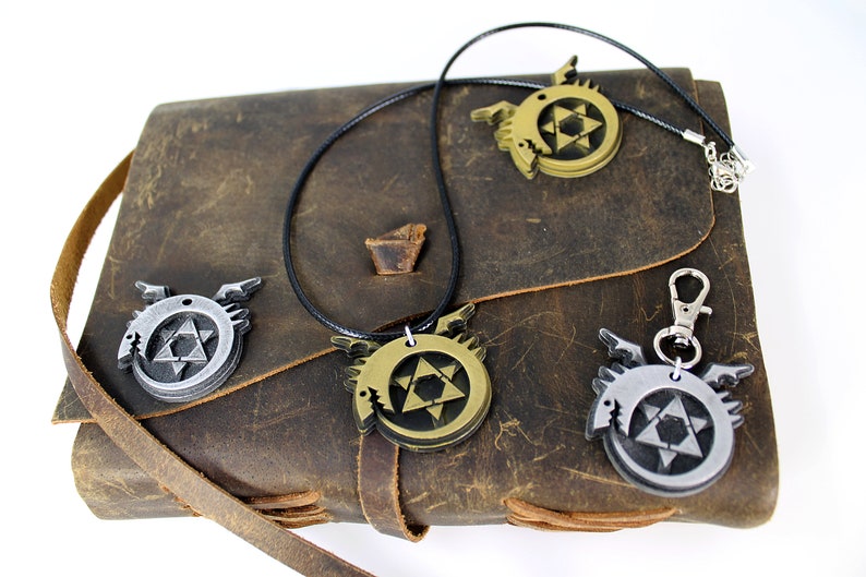 May include: Three metal pendants featuring a stylized winged serpent with a star of David inside. The pendants are in silver, gold, and bronze and are displayed on a brown leather journal.