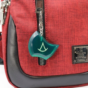 May include: A red crossbody bag with a black zipper and a green plastic charm with a white symbol. The bag has a black leather trim and a silver metal tag that reads "Where is the heart to receive good wishes".