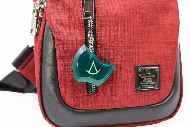 May include: A red crossbody bag with a black zipper and a green plastic charm with a white symbol. The bag has a black leather trim and a silver metal tag that reads "Where is the heart to receive good wishes".