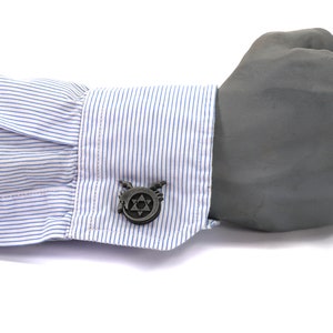 May include: A pair of silver cufflinks with a black and white design of a star with wings. The cufflinks are on a blue and white striped shirt cuff.
