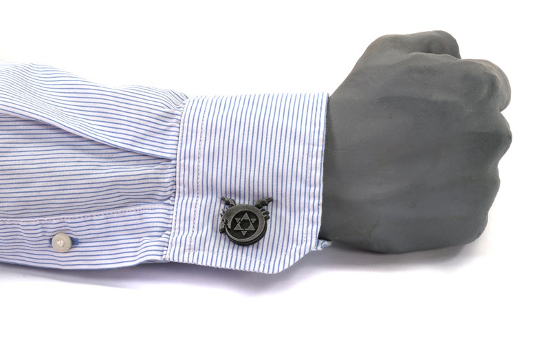 May include: A pair of silver cufflinks with a black and white design of a star with wings. The cufflinks are on a blue and white striped shirt cuff.