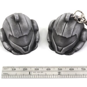 Puede incluir: Dos llaveros de metal de color plateado con forma de casco. El llavero de la derecha tiene un anillo de metal plateado unido a &eacute;l. Una regla est&aacute; en la parte inferior de la imagen con marcas en pulgadas.