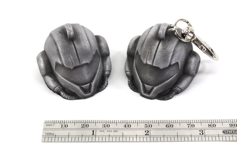 Puede incluir: Dos llaveros de metal de color plateado con forma de casco. El llavero de la derecha tiene un anillo de metal plateado unido a &eacute;l. Una regla est&aacute; en la parte inferior de la imagen con marcas en pulgadas.
