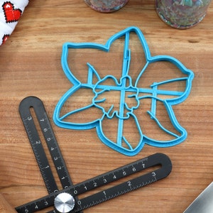 May include: A blue plastic cookie cutter shaped like a daffodil flower. The cutter is on a wooden surface with a black ruler and a rolling pin.