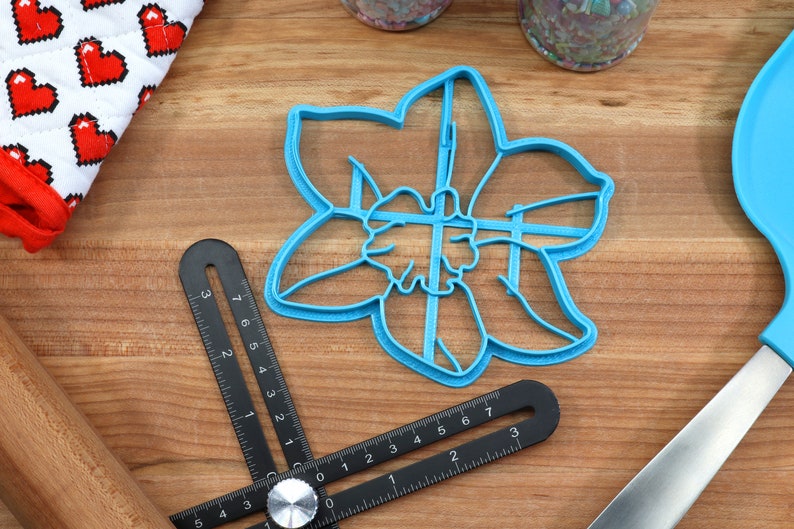 May include: A blue plastic cookie cutter shaped like a daffodil flower. The cutter is on a wooden surface with a black ruler and a rolling pin.