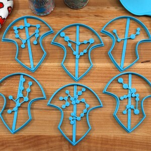 Puede incluir: Seis cortadores de galletas de pl&aacute;stico azul en forma de escudos con dise&ntilde;os de constelaciones en el interior. Los cortadores est&aacute;n sobre una superficie de madera.
