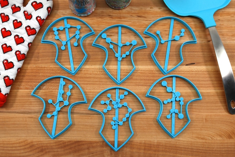 Puede incluir: Seis cortadores de galletas de pl&aacute;stico azul en forma de escudos con dise&ntilde;os de constelaciones en el interior. Los cortadores est&aacute;n sobre una superficie de madera.