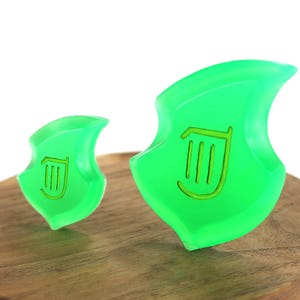 May include: Two green plastic charms shaped like a leaf with a harp symbol in the center. The charms are sitting on a wooden surface.