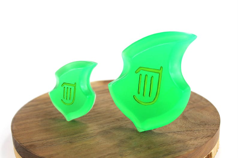 May include: Two green plastic charms shaped like a leaf with a harp symbol in the center. The charms are sitting on a wooden surface.