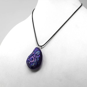 Puede incluir: Un collar con un colgante de vidrio iridiscente morado y rosa con un s&iacute;mbolo rosa en un cord&oacute;n negro.
