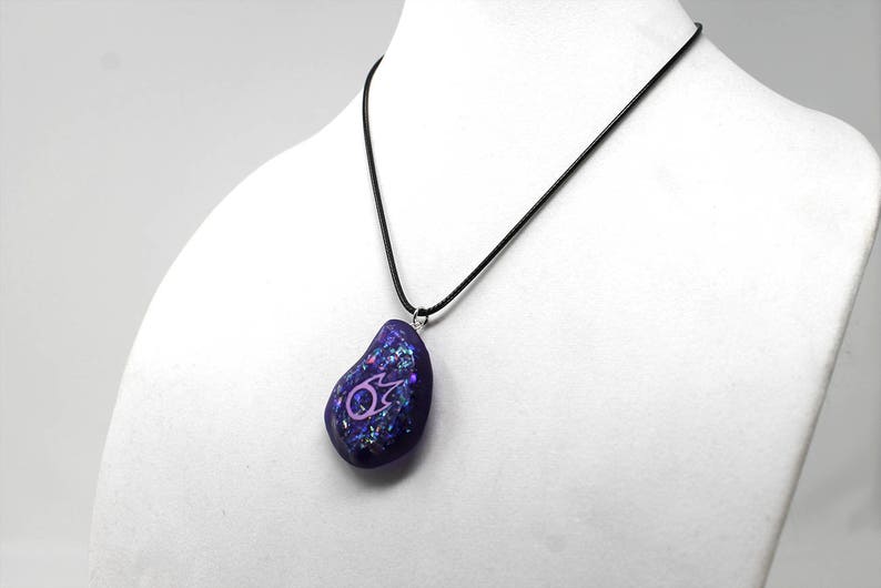 Puede incluir: Un collar con un colgante de vidrio iridiscente morado y rosa con un s&iacute;mbolo rosa en un cord&oacute;n negro.