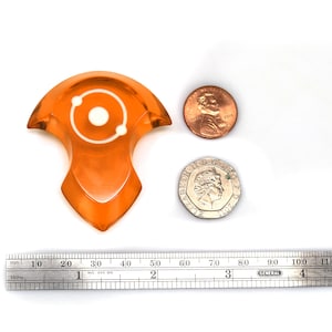 May include: Orange resin tabletop dice with a white atom symbol. The dice is about 2 inches tall and is next to a US penny and a British coin for scale.