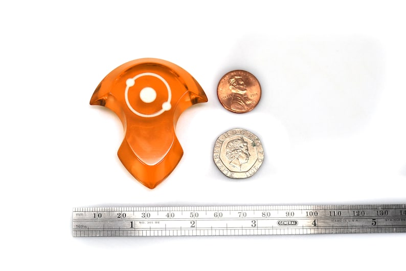 May include: Orange resin tabletop dice with a white atom symbol. The dice is about 2 inches tall and is next to a US penny and a British coin for scale.