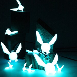May include: A collection of glowing blue fairy keychains with wings. The keychains are arranged on a dark surface with a few on wooden blocks.
