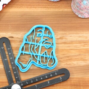 May include: A blue plastic cookie cutter shaped like a dog's head. The cutter is on a wooden surface with a ruler and a jar of sprinkles.