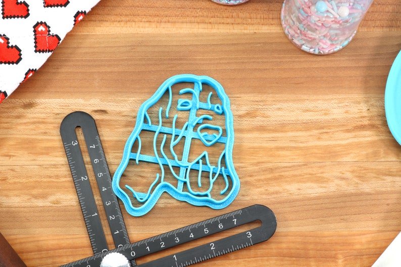 May include: A blue plastic cookie cutter shaped like a dog's head. The cutter is on a wooden surface with a ruler and a jar of sprinkles.