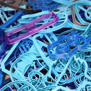 Op de afbeelding: Een verzameling blauwe, paarse en grijze plastic koekjesvormen in verschillende vormen. De vormen zijn in een stapel gerangschikt, waarbij sommige elkaar overlappen.