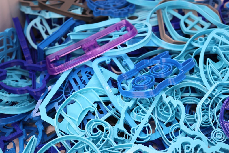 Op de afbeelding: Een verzameling blauwe, paarse en grijze plastic koekjesvormen in verschillende vormen. De vormen zijn in een stapel gerangschikt, waarbij sommige elkaar overlappen.
