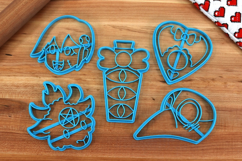 Pode incluir: Cinco cortadores de biscoitos de pl&aacute;stico azul em forma de cora&ccedil;&atilde;o com um la&ccedil;o, uma garrafa com olhos, um personagem com chifres e um pentagrama, um personagem com um curativo e um personagem com um bico.