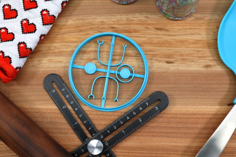 May include: A blue plastic cookie cutter shaped like a circle with a design inside. The design includes two circles, two curved lines, and two small loops.