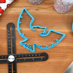 May include: A blue plastic bat-shaped cookie cutter, a black multi-angle measuring tool, and a blue spatula are on a wooden surface. A white cloth with red pixelated hearts and jars of sprinkles are also present. Baking and crafting supplies.