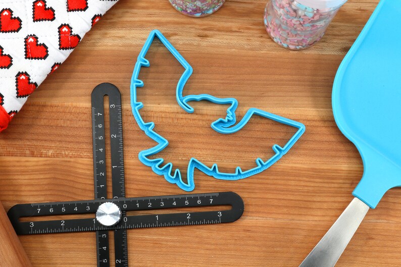 May include: A blue plastic bat-shaped cookie cutter, a black multi-angle measuring tool, and a blue spatula are on a wooden surface. A white cloth with red pixelated hearts and jars of sprinkles are also present. Baking and crafting supplies.