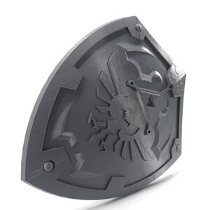 May include: A gray plastic shield with a detailed design of a stylized bird with wings spread, a triangle, and other geometric shapes. The shield has a textured surface and appears to be a prop or replica.