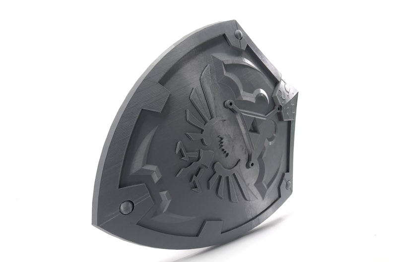 May include: A gray plastic shield with a detailed design of a stylized bird with wings spread, a triangle, and other geometric shapes. The shield has a textured surface and appears to be a prop or replica.
