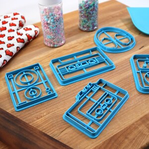 May include: A set of five blue plastic cookie cutters shaped like a record player, a cassette tape, and a record. The cutters are on a wooden surface.