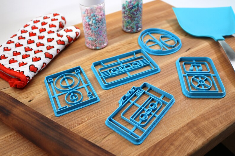 May include: A set of five blue plastic cookie cutters shaped like a record player, a cassette tape, and a record. The cutters are on a wooden surface.