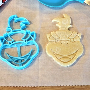 May include: A blue plastic cookie cutter and a yellow cookie in the shape of a cartoon character's head. The cookie cutter and cookie are on a white surface.