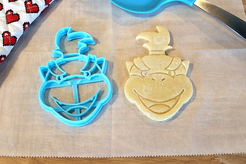 May include: A blue plastic cookie cutter and a yellow cookie in the shape of a cartoon character's head. The cookie cutter and cookie are on a white surface.