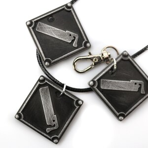 May include: A silver necklace with three square pendants featuring a saw blade design. The necklace has a black cord and a silver clasp.