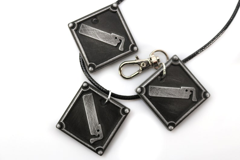 May include: A silver necklace with three square pendants featuring a saw blade design. The necklace has a black cord and a silver clasp.