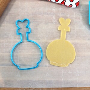 May include: A blue plastic cookie cutter shaped like a bottle with a heart-shaped top, and a yellow cookie dough cut out in the same shape.