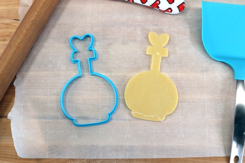 May include: A blue plastic cookie cutter shaped like a bottle with a heart-shaped top, and a yellow cookie dough cut out in the same shape.