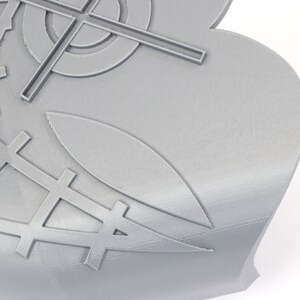 May include: A close-up of a gray, 3D-printed object with a complex geometric design. The design includes crosshairs, a grid pattern, and stylized leaf shapes. The object has a smooth, matte finish and is set against a white background.