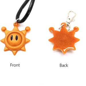 May include: A gold-colored sun pendant with a black face and two eyes. The pendant is attached to a black cord necklace. The back of the pendant is plain gold.