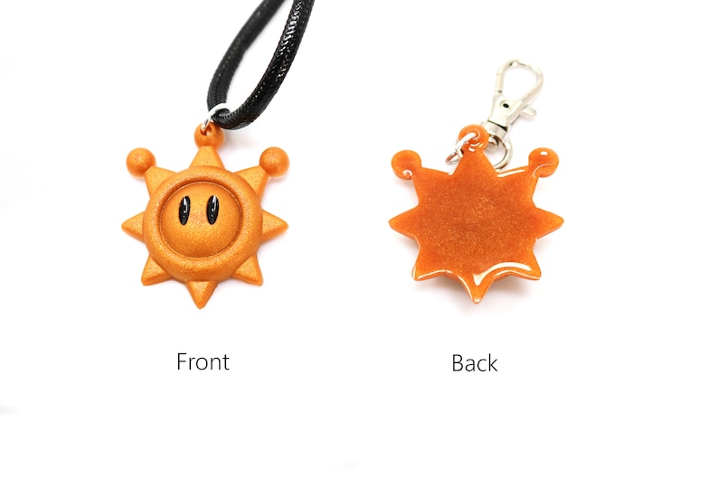 May include: A gold-colored sun pendant with a black face and two eyes. The pendant is attached to a black cord necklace. The back of the pendant is plain gold.