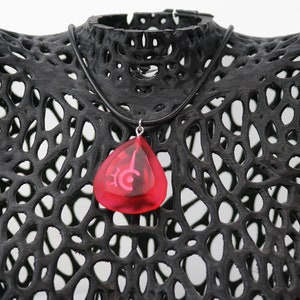 May include: A red teardrop-shaped pendant necklace with a silver chain and a black cord. The pendant has a white symbol inside.