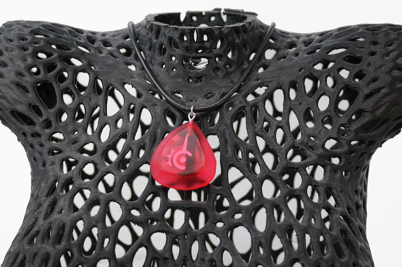 May include: A red teardrop-shaped pendant necklace with a silver chain and a black cord. The pendant has a white symbol inside.