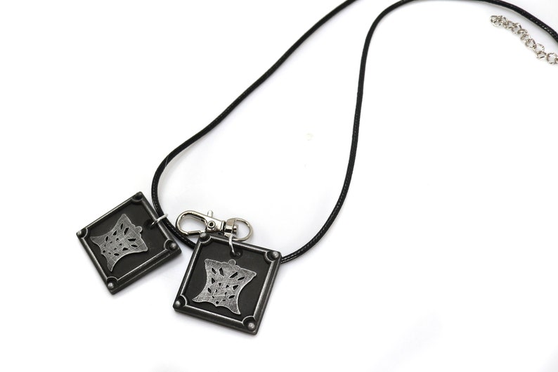 Puede incluir: Un collar de cord&oacute;n negro con un colgante de metal plateado que presenta un dise&ntilde;o detallado. El colgante est&aacute; unido a un cierre de metal plateado.