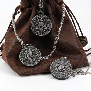 May include: Three silver-colored pendant necklaces with a circular design and chain are displayed in front of a brown leather pouch. The pendants have an engraved star and text. The pouch is textured and has a drawstring closure.