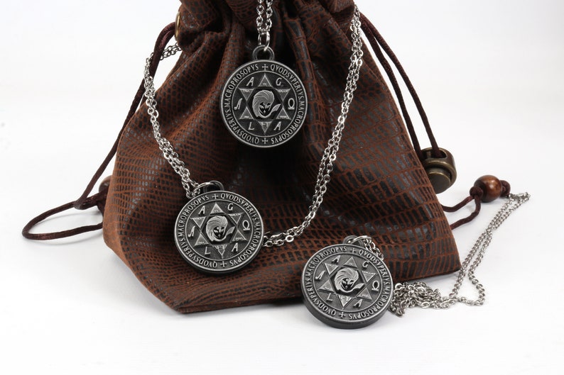 May include: Three silver-colored pendant necklaces with a circular design and chain are displayed in front of a brown leather pouch. The pendants have an engraved star and text. The pouch is textured and has a drawstring closure.