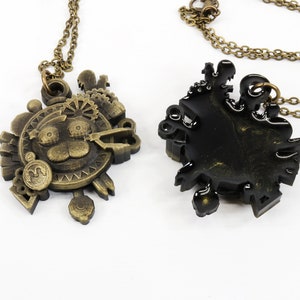 May include: Two gold-tone necklaces with intricate, detailed pendants. One pendant features a cartoon animal face with a gear design, the other is a more abstract design. Both pendants are on a chain.