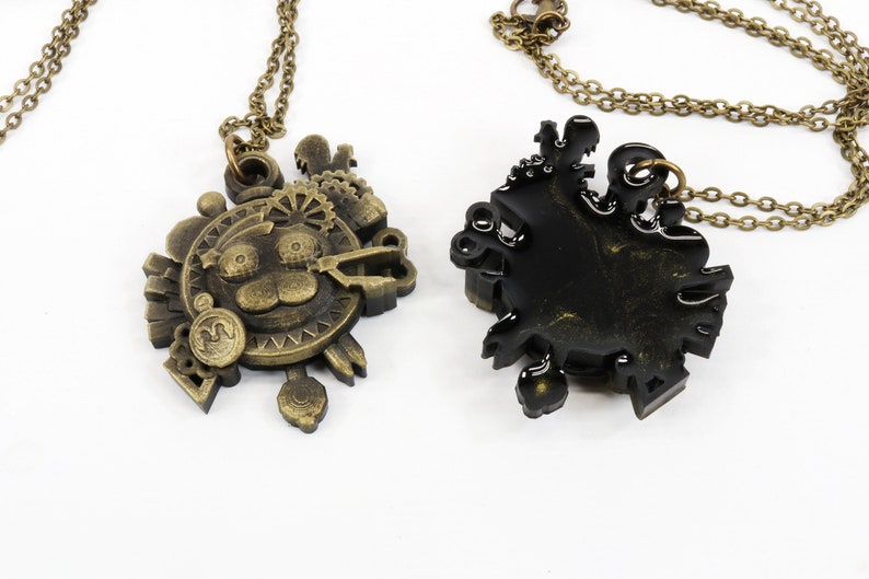 May include: Two gold-tone necklaces with intricate, detailed pendants. One pendant features a cartoon animal face with a gear design, the other is a more abstract design. Both pendants are on a chain.