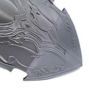 May include: A gray, intricately detailed shield with a pointed bottom and a raised, decorative design. The shield is made of a smooth, hard material.