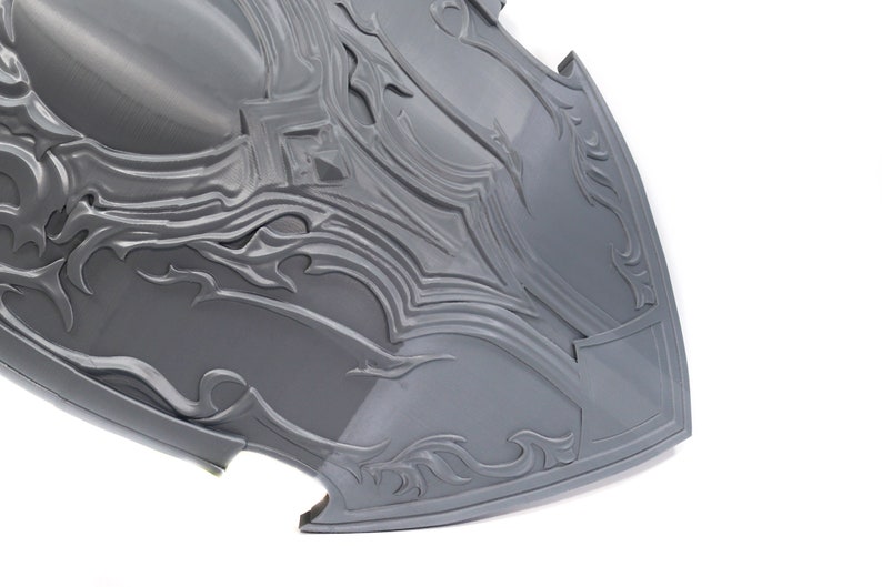 May include: A gray, intricately detailed shield with a pointed bottom and a raised, decorative design. The shield is made of a smooth, hard material.