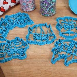 May include: Five blue plastic cookie cutters shaped like cartoon faces. The cutters are on a wooden surface.