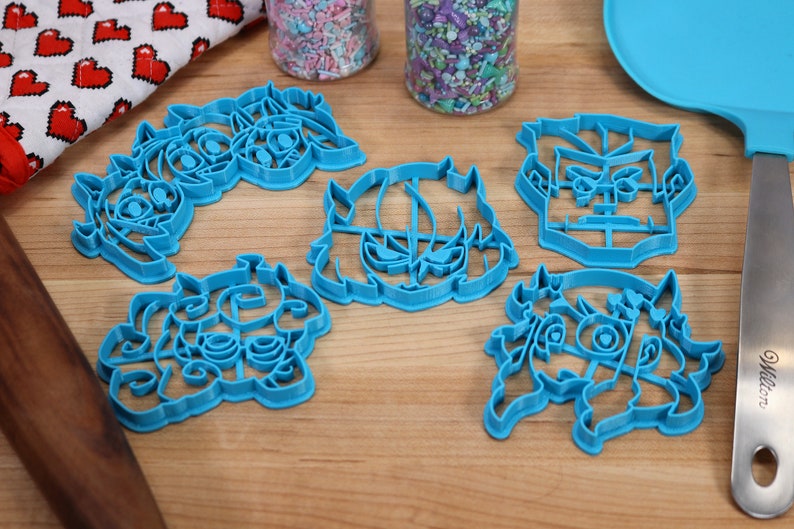 May include: Five blue plastic cookie cutters shaped like cartoon faces. The cutters are on a wooden surface.